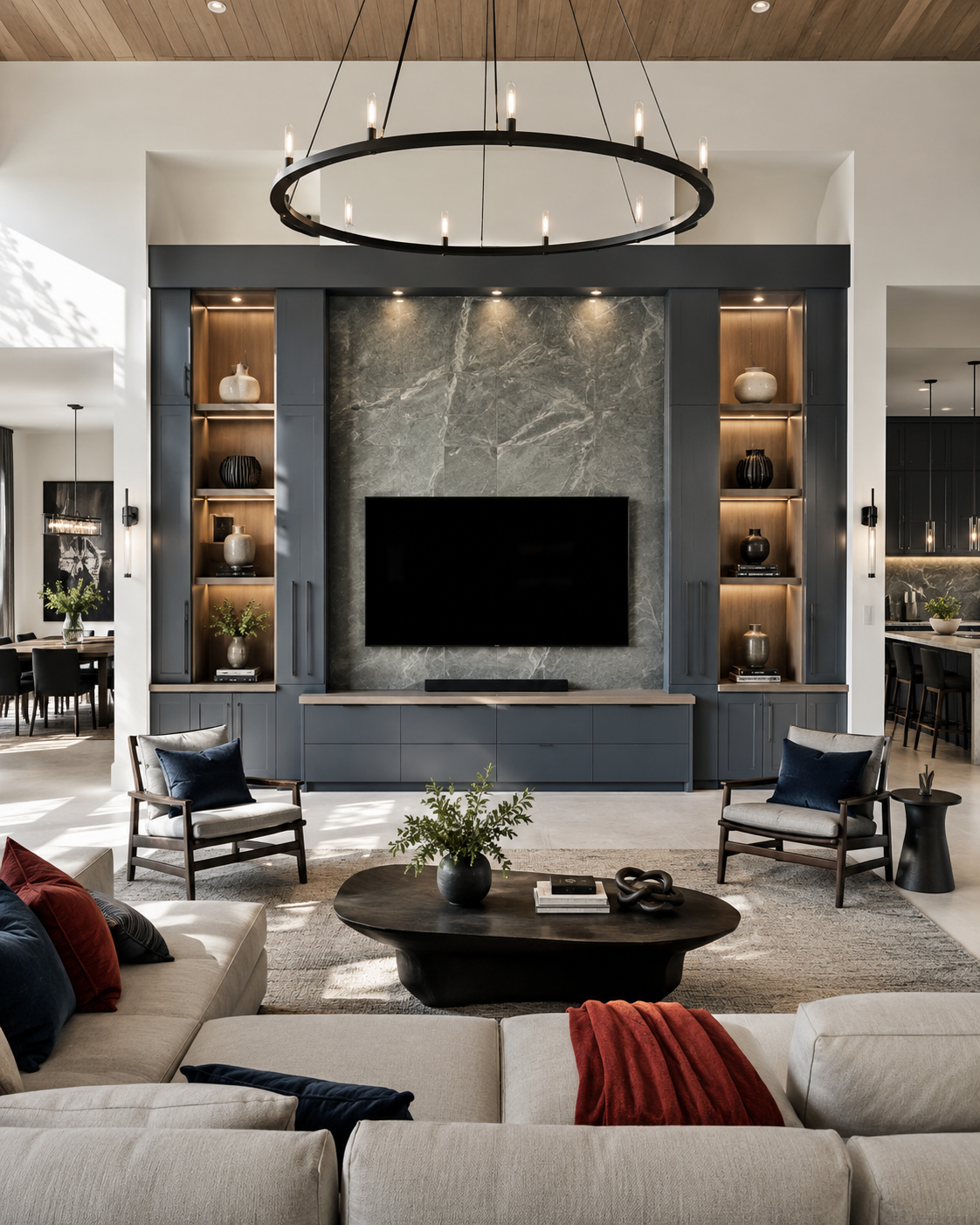 Large open-concept great room with a tall gray stone-look TV wall, charcoal blue built-ins, illuminated wood shelving, lower cabinets, high ceilings, and views into adjacent dining and kitchen areas.