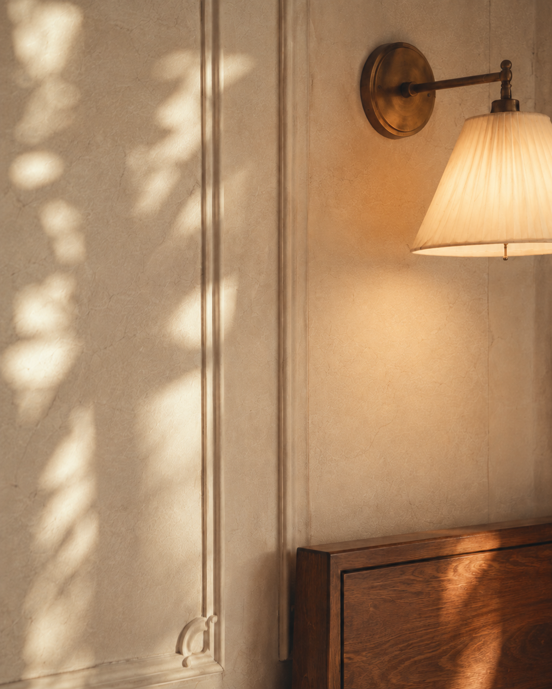 Long ivory curtain moving in an open-window breeze while sunlight and a glowing wall sconce shape a paneled bedroom wall.