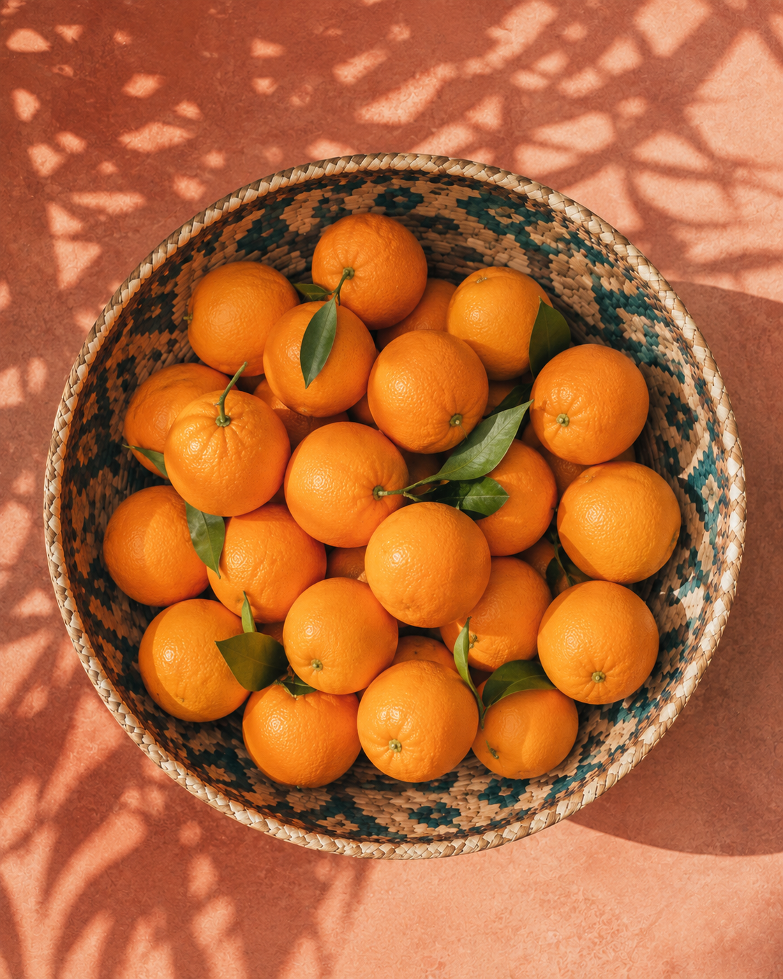 Top-down view of bright Florida oranges in a woven basket on a warm terracotta surface with sunlit leaf shadows.