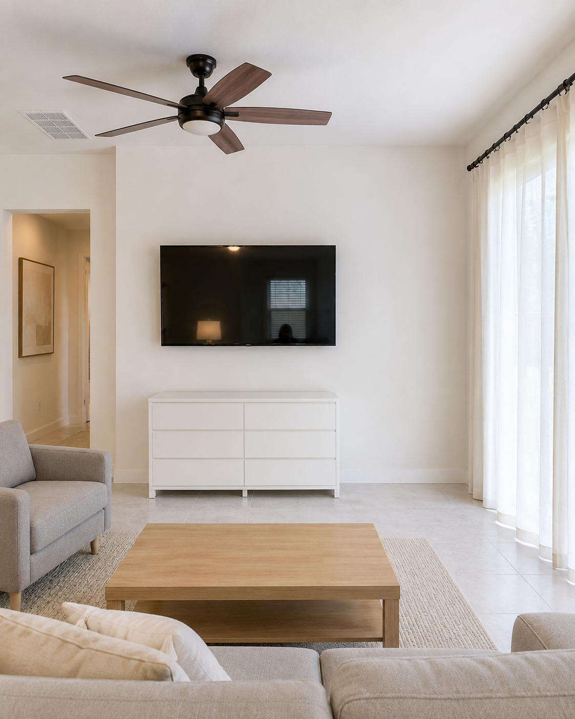 Before view of the Sarasota living room showing a TV mounted on a plain wall above a freestanding white dresser, with an interior doorway on the left and a large sliding glass door on the right.