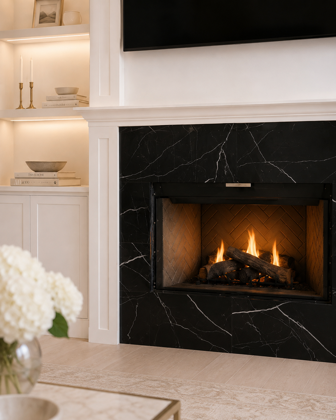 Detail view of the Windermere fireplace showing the black marble-look surround with white veining, traditional firebox, classical white mantel trim, and warm illuminated built-in shelving beside it.