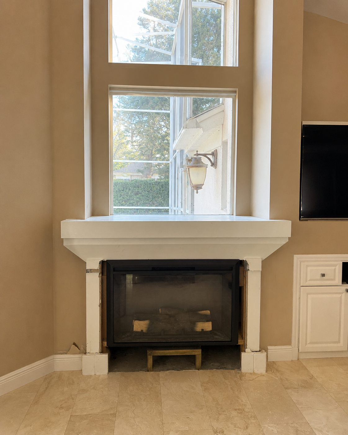 Before view of the Winter Park living room showing the existing fireplace beneath a tall window, unfinished and dated surround trim, and the adjacent built-in cabinet and wall-mounted TV before renovation.