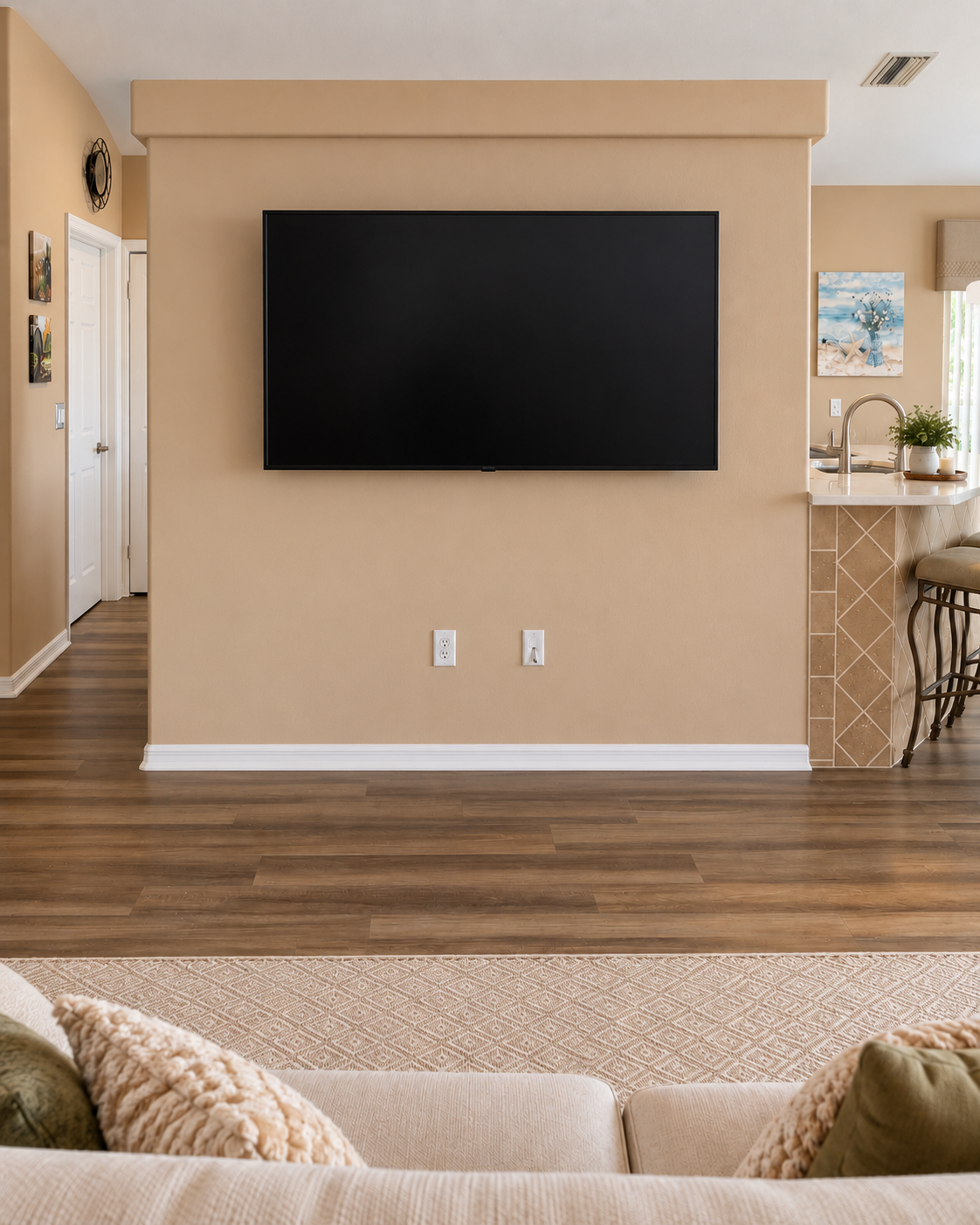 Before photograph of the original beige great room wall with a mounted television and two outlets below, prior to the fireplace and mantel installation.
